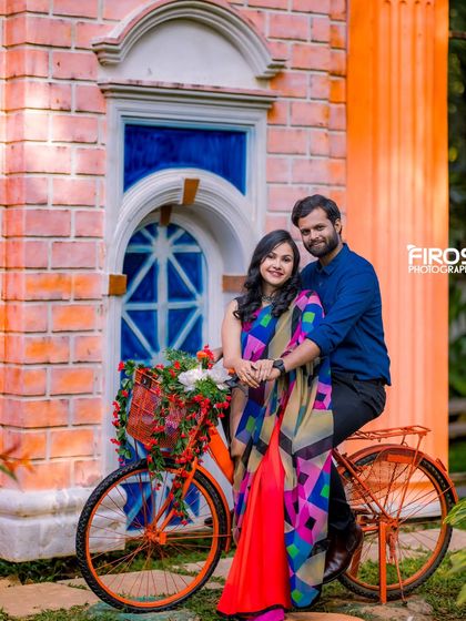 A couple poses with a decorated orange bicycle in front of a colorful building, surrounded by the greenery of the garden.