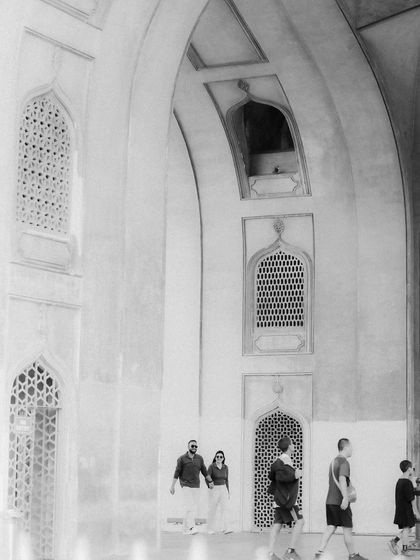 A wide, atmospheric shot in black and white, showing the couple walking through a grand hall with intricate archways. The presence of other people adds a sense of real-world context to their story.