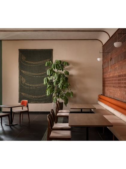 A seating arrangement at Ishtaa featuring a mix of chair styles and a large piece of textile art. The placement of greenery and the warm terracotta brick wall create a comfortable and visually interesting corner for diners.