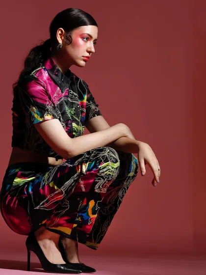 A model in a vibrant, abstract-print co-ord set. The crouching pose and intense gaze, combined with the bold pink background, create a powerful and modern fashion statement.