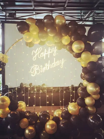 A chic and modern birthday setup using a black and gold balloon ring against a simple white background with fairy lights.