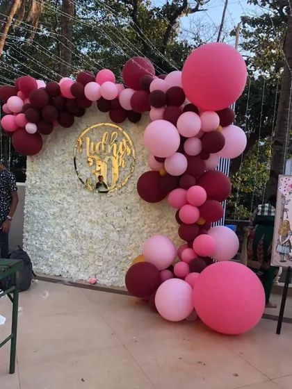 An outdoor balloon arch against a white floral wall. The organic garland in shades of pink and maroon with a custom gold nameplate creates a beautiful photo opportunity.