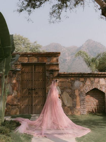 A breathtaking shot of the bride against a rustic stone wall and mountains. The incredibly long, sheer pink veil creates a dramatic and ethereal effect, making for a truly memorable bridal portrait.