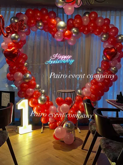 A classic first-anniversary decoration in a restaurant. This setup includes a large heart-shaped balloon arch in red and silver, a custom "Happy Anniversary" neon sign, and a light-up marquee number "1".