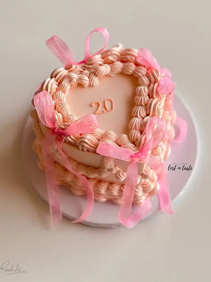 A beautiful heart-shaped cake in a soft pink hue, decorated with classic vintage Lambeth piping and delicate organza ribbons. This design is perfect for celebrating a 20th birthday in style.