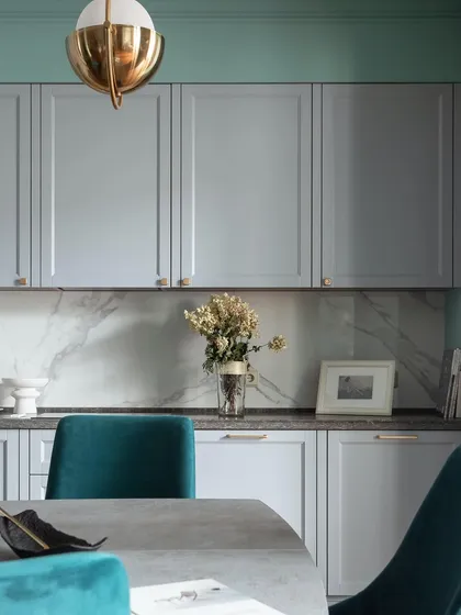 A sophisticated kitchen and dining area with PU-finished grey cabinets, a marble backsplash, and plush teal dining chairs.