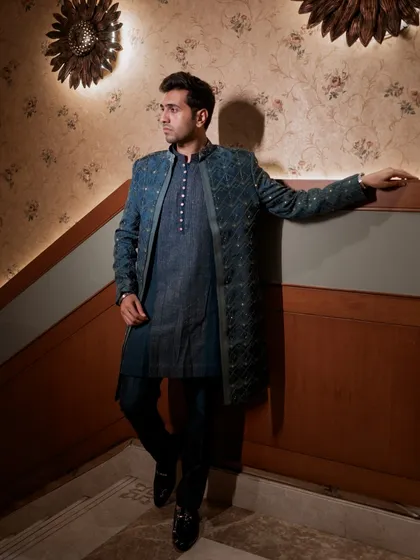 A classic groom portrait against a patterned wall. His pose is relaxed but poised, and the warm lighting complements his deep teal outfit.