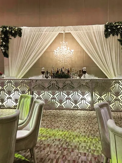 An elegant bar setup for a Roka ceremony, featuring a backlit geometric-patterned bar front, a crystal chandelier, and a backdrop of white drapes.