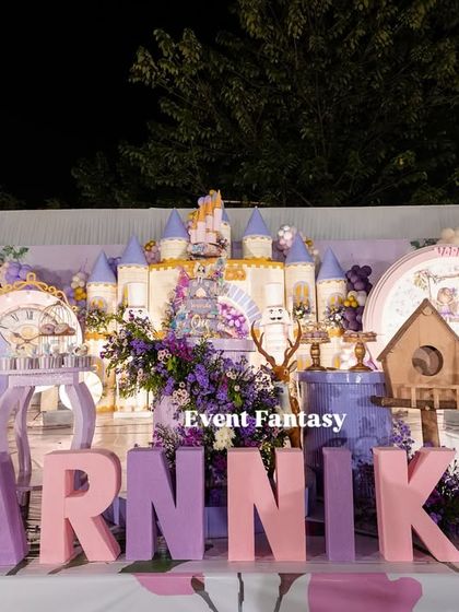 A full stage view of Varnnika's fairyland, a stunning setup in shades of purple and pink. It features a large castle backdrop, oversized name blocks, fairy dolls, and whimsical butterfly wing props.