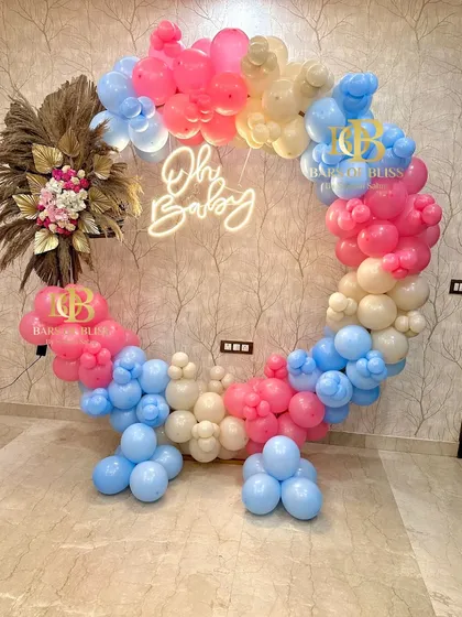 A colorful 'Oh Baby' gender reveal or baby shower setup. The full balloon ring incorporates pink, blue, and cream balloons, with a floral pampas grass accent and a central neon sign.