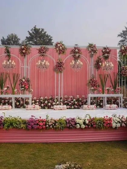 A beautiful outdoor stage with a pink backdrop and white metal arches. The arches are decorated with small chandeliers and floral arrangements, and the stage is lined with a dense border of pink, white, and purple flowers.