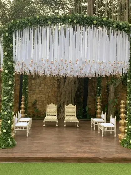 A round mandap design framed with lush greenery and white ribbon details. The use of traditional golden pillars adds a classic touch to this modern and elegant structure.
