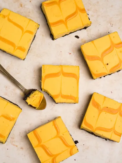 A flatlay of mango cheesecake bars, shot for a dessert brand. The geometric arrangement and the single bite taken out with a spoon make the image visually interesting and tempting.