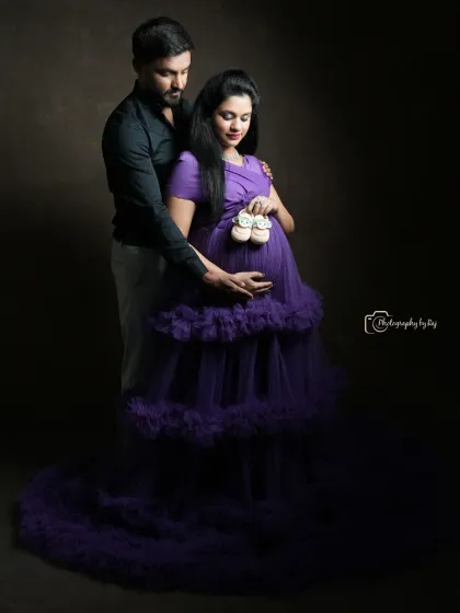 A tender studio portrait of a couple holding tiny baby shoes over the bump. The mother-to-be is wearing a beautiful purple ruffled gown.