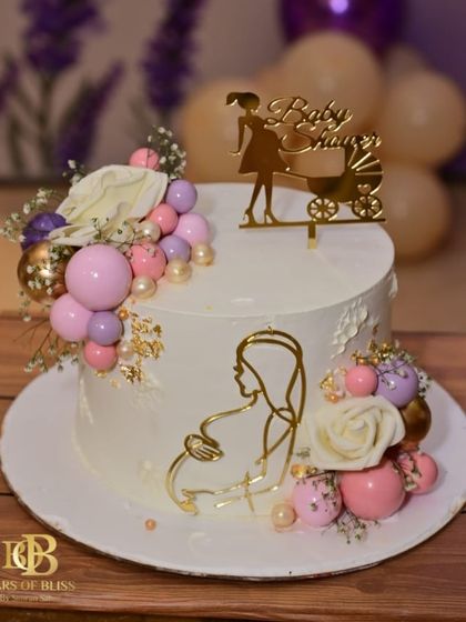 A beautiful and elegant cake to celebrate a baby shower. This design features a gold acrylic silhouette of a mother-to-be, a "Baby Shower" topper, and a cascade of pink, purple, and gold spheres.