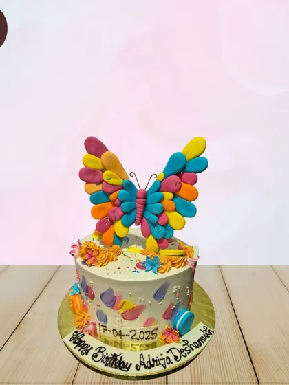 A simple and colorful butterfly cake. The main feature is a large, multi-colored butterfly topper made of individual, vibrant petals.