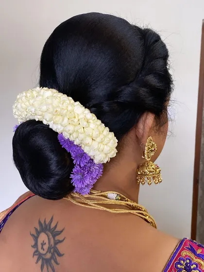 A classic bridal bun adorned with a jasmine gajra and a pop of purple flowers. The tattoo on the bride's neck adds a modern and personal touch to the traditional look.