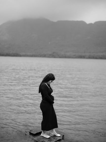 A dramatic black and white portrait of the mom-to-be standing on a rock by the water, with mountains in the background. It’s a powerful image of motherhood and nature.