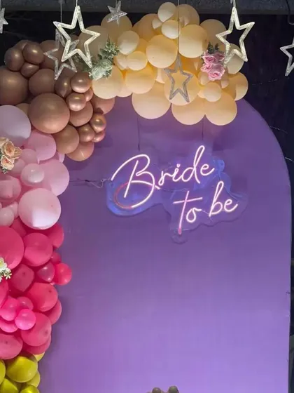A stylish and colorful photo booth featuring a purple arched panel and a "Bride to be" neon sign. The organic balloon garland in shades of pink, yellow, and brown adds a playful and modern touch to the setup.