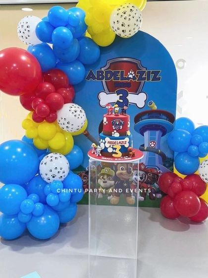 A vibrant Paw Patrol themed third birthday setup. The decoration includes a balloon arch in the classic red, blue, and yellow colors, with a cake placed on a clear pedestal.