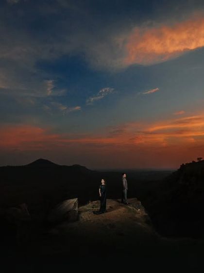 A dramatic wide shot of a couple standing on a cliff edge against a colorful sunset sky. The lighting is carefully crafted to highlight them amidst the vast, dark landscape.