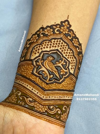 A close-up of a delicate arm band featuring a bird perched on a branch inside a decorative frame. The fine lines and shading give the design a beautiful depth.