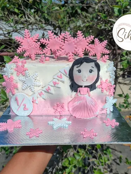 A sweet six-month milestone celebration cake. This half-birthday cake is decorated with pink and white snowflakes and a cute figurine of the birthday girl.