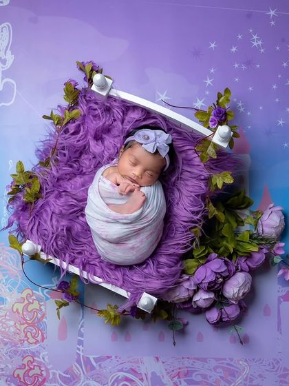An artistic shot of a newborn in a white prop bed, set against a dreamy purple digital backdrop with stars and castle illustrations.