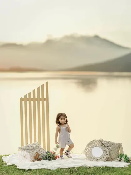 A toddler stands on a picnic blanket by a lake at sunset. The warm light, calm water, and distant mountains create a stunning backdrop for this beautiful and peaceful milestone portrait.
