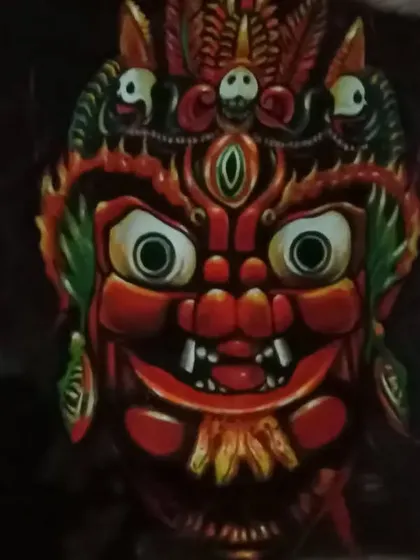 A painting of a red-faced deity mask with wide eyes and intricate headgear. The dark background makes the vibrant colors and expressive features of the mask stand out.