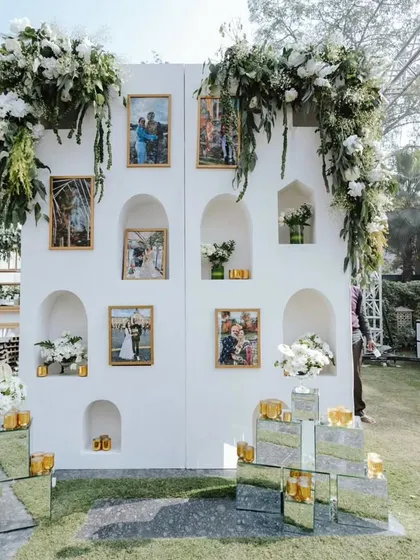 A personalized memory wall we created for a garden wedding. The white structure with arched niches displayed the couple's photos, while fresh white and green floral garlands added a touch of natural elegance.