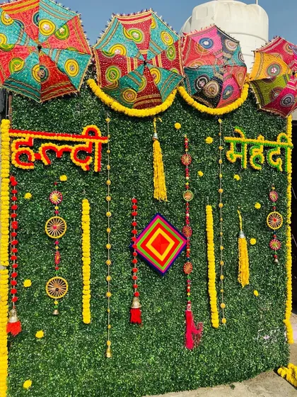 For a modern take on traditional Haldi and Mehndi decor, I use an artificial green wall as a base. This backdrop is decorated with marigold garlands, colorful tassels, and Rajasthani umbrellas to create a vibrant and festive photo corner.