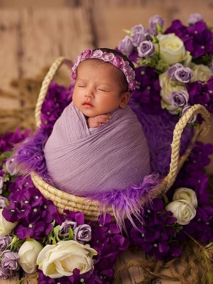 This newborn is peacefully asleep in a basket overflowing with purple and white flowers. The textures of the feathers, flowers, and wrap create a visually stunning composition.