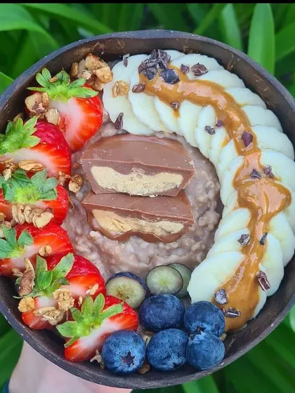 A delicious and healthy breakfast bowl with oatmeal, banana, strawberries, blueberries, and a peanut butter chocolate bar.