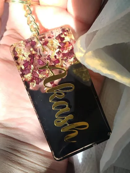 This keychain is a beautiful way to preserve a memory. I used crushed rose petals and gold flakes at the top of this rectangular keychain, with the name 'Akash' in a flowing gold script on a solid black background.