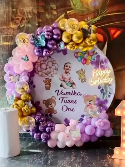 A sweet teddy bear theme for Vamika's first birthday. The circular backdrop features a personalized print and is surrounded by a purple and pink balloon garland with butterfly and bear details.
