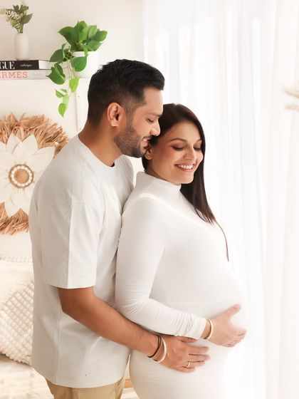 A classic and elegant pose by the window. The expecting couple is bathed in soft, natural light, highlighting the baby bump and their peaceful connection.