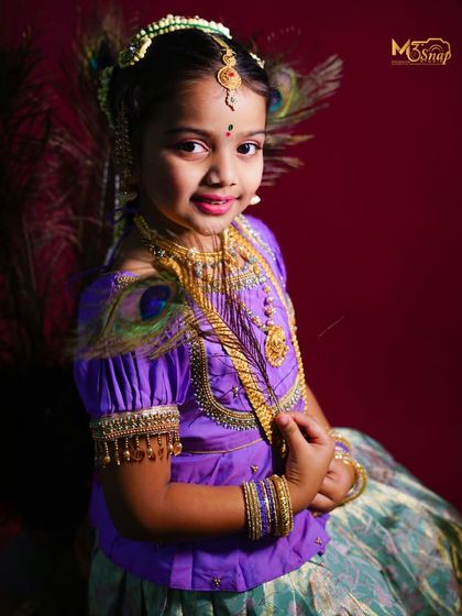 A close-up portrait against a rich, dark red background. The lighting highlights the intricate details of her jewelry and the textures of the peacock feathers.