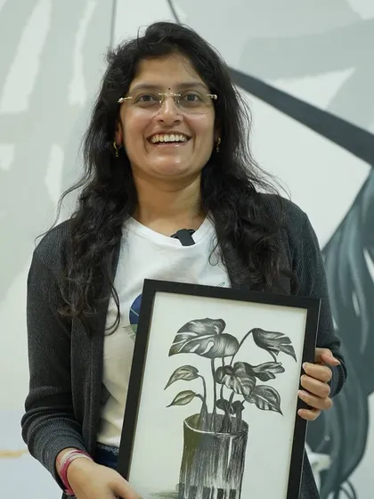 A participant proudly displays her framed charcoal drawing of a monstera plant. This workshop focuses on shading and texture to create realistic black and white art.