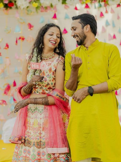 A sweet moment from the Mehendi ceremony, where the couple laughs together while the bride holds a "His" prop. It’s a perfect capture of their playful dynamic and the fun of the event.