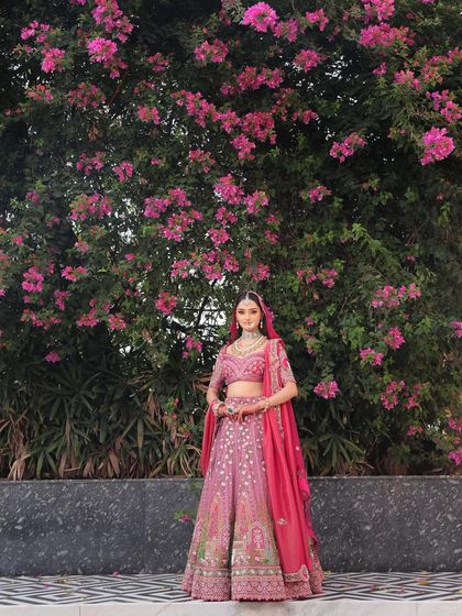 A full-length shot showing the bride's stunning pink lehenga. The soft, glowing makeup complements the vibrant outfit perfectly.