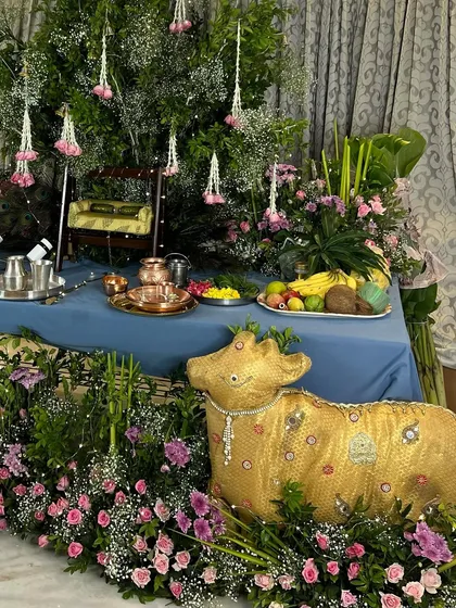 A wider view of the Janmashtami setup, showing the golden cow prop in context. The altar is adorned with fresh fruit, copper utensils, and a miniature swing, all surrounded by abundant flowers and greenery.