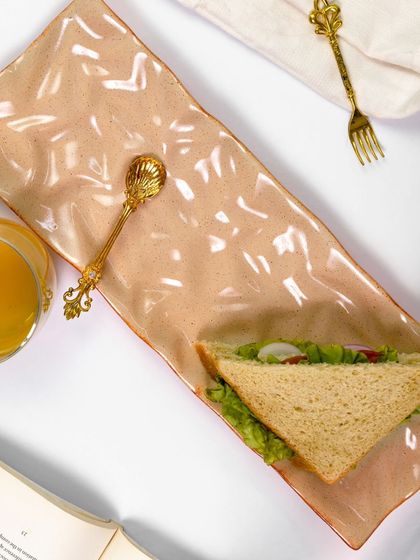 A top-down view of the long ceramic platter, showcasing its unique texture and shape. The simple styling with a sandwich and golden cutlery creates a balanced and aesthetically pleasing flatlay.