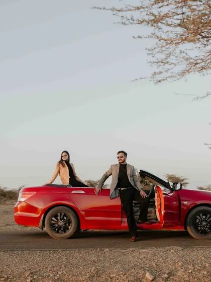 A wide shot of the couple with their convertible on a desert road, framed by a tree, creating a cinematic, journey-like feel.