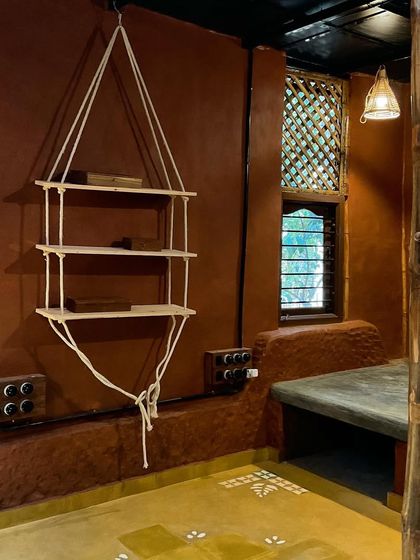 The interior of a mud house with wattle and daub walls, built-in bamboo furniture, and exposed electrical conduits for a rustic, honest aesthetic.