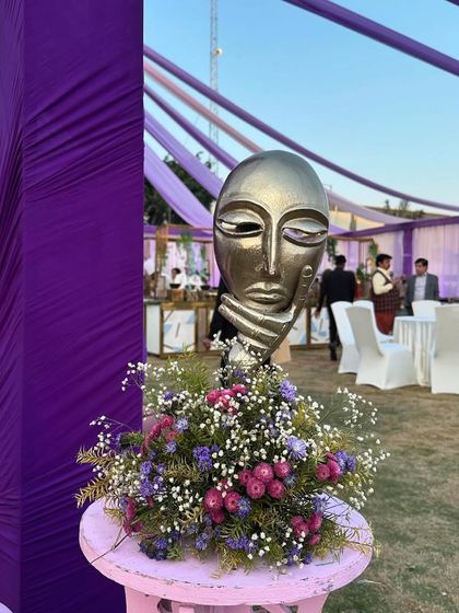 An artistic centerpiece with a golden mask sculpture and fresh flowers, adding a touch of mystique to the Haldi carnival theme.
