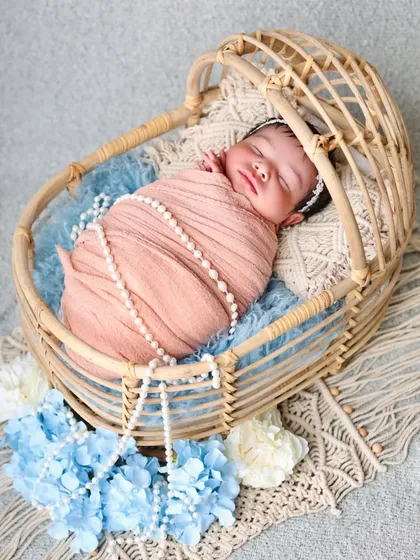 A newborn girl wrapped in a soft peach swaddle with a pearl necklace, sleeping in a beautiful cane bassinet. The blue and white flowers add a lovely color contrast.