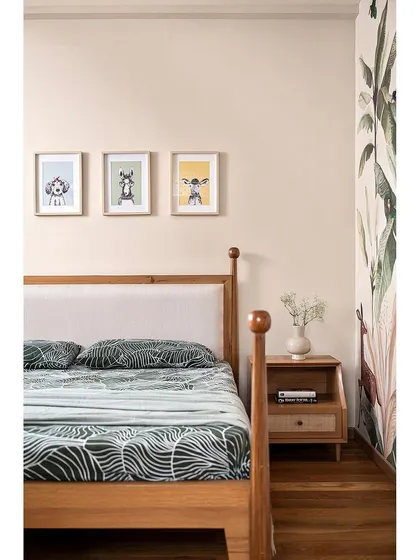 A close-up of the bed in the jungle-themed room. The simple wooden frame is set against the vibrant wallpaper, and cute animal-themed art prints add to the playful atmosphere.