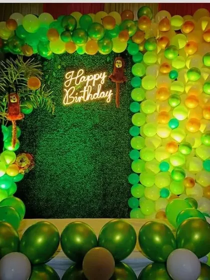 A jungle-themed birthday backdrop for an outdoor or semi-outdoor space. The artificial grass wall is framed by a green and yellow balloon arch and lit by a neon sign.