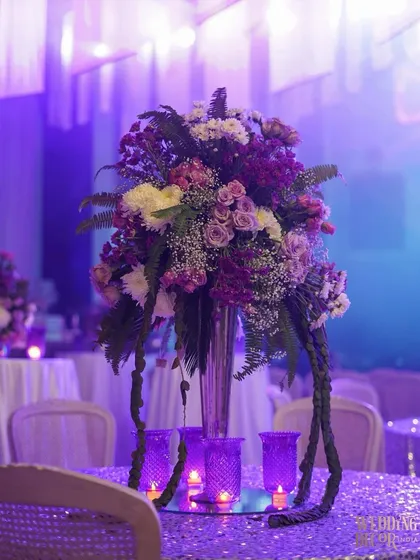 A beautiful table centerpiece featuring a tall floral arrangement in a silver vase, surrounded by purple candle holders. The sequined tablecloth adds a touch of sparkle to the setting.